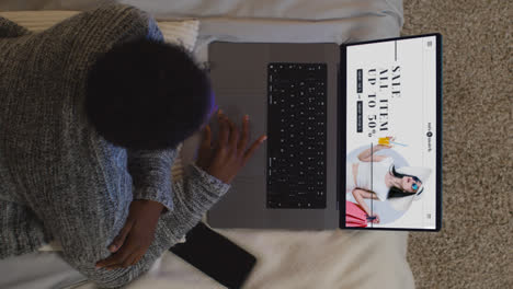 Overhead-Shot-Of-Woman-Lying-On-Bed-At-Home-Looking-At-Online-Fashion-Sale-Bargains-On-Laptop